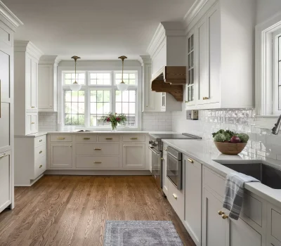 Elegant kitchen with white cabinets, brass handles, wooden floor, and large windows. A bowl of vegetables and flowers adorns the countertops.