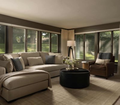 A cozy living room with beige sofa, patterned cushions, a round black coffee table, and large windows overlooking a sunny green garden.