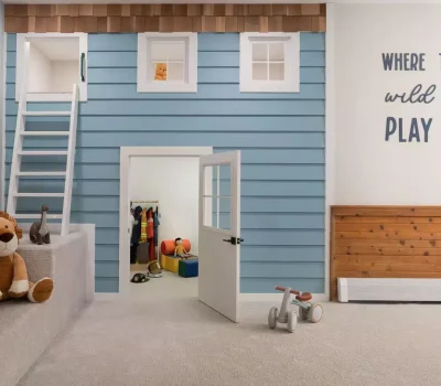 Playroom with blue playhouse, ladder, and stuffed animals. Wall reads "Where the Wild Ones Play." Toys and costumes inside. Cozy, welcoming atmosphere.