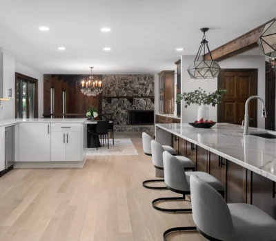 Modern kitchen with white cabinets, marble countertops, large island, geometric pendant lights, and stone fireplace in connected dining area, featuring wood accents and greenery.