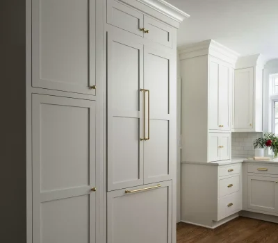 A modern kitchen with white cabinets, gold handles, and wooden floors. Natural light illuminates the space, highlighting a vase of flowers.