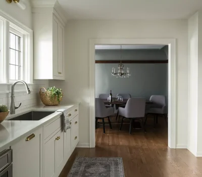 Elegant kitchen with white cabinets, bowl of vegetables, and a sink. Leads to a dining area with a chandelier and gray chairs.