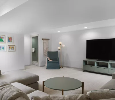 A modern living room features a sectional sofa, large TV, artwork on walls, cozy armchair, round coffee table, and soft ambient lighting.