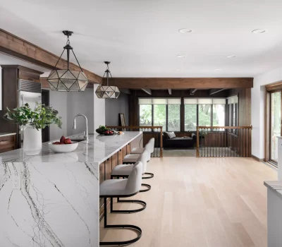 Modern kitchen with marble countertops, geometric light fixtures, wooden beams, and large windows overlooking greenery. Includes a spacious island and seating area.