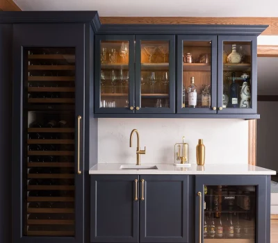 A home bar with dark cabinets, featuring a wine fridge, glass storage, golden faucet, and countertop, set against light wooden flooring and beams.