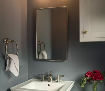 A modern bathroom features a white pedestal sink, rectangular mirror, wall light, and a vase with red flowers near the toilet.