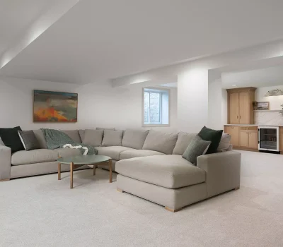 A modern basement features a large sectional sofa, abstract artwork, beige carpeting, and a wet bar with light wood cabinetry and a mini-fridge.