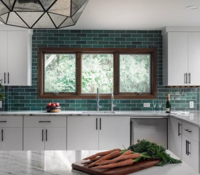 Modern kitchen with white cabinets, green subway tile backsplash, geometric pendant light, and counter with fresh carrots and apples. Large window overlooks greenery.