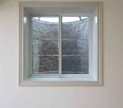 A closed window in a room reveals a stone window well with a mesh cover, providing limited natural light and view.