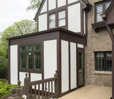 Tudor-style house with timber detailing, adjacent brick section, and a wooden ramp. Surrounded by greenery under a cloudy sky.