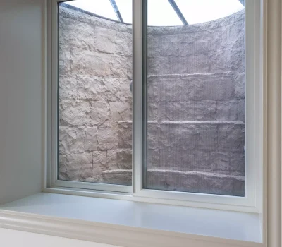 Basement window view showing an enclosed window well with a stone wall and a metal grate cover, allowing limited light into the space.