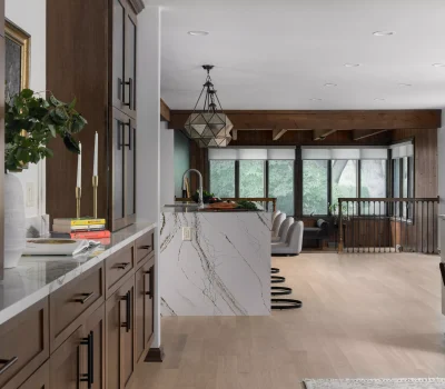 Modern, open-plan kitchen and living area with wooden cabinetry, marble countertops, geometric pendant light, and large windows overlooking greenery. Minimalist decor and natural light.