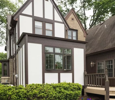 Tudor-style house with wood trim, brick accents, porch, and vibrant garden. Surrounded by lush green trees, overcast sky backdrop.