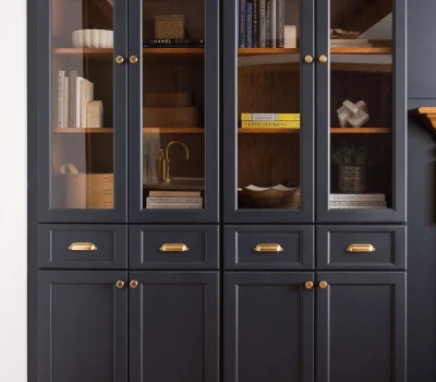 A black cabinet with glass doors displays books, ceramics, and decorative items. It has gold handles and wooden shelves, against a wooden floor.