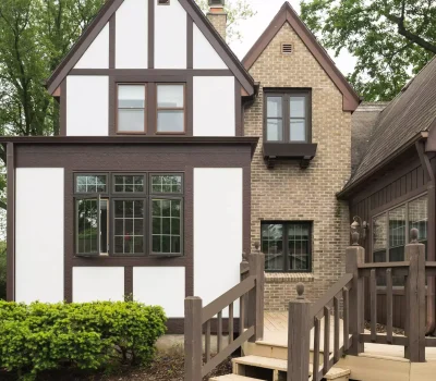 A charming Tudor-style house with brick and timber elements, surrounded by lush greenery and colorful flowers, features a small wooden staircase.