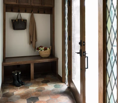 A cozy entryway with a wooden door, hexagonal tile floor, and bench. Contains boots, coat, handbag, and a basket of flowers.
