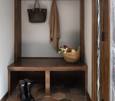 Mudroom with wooden bench and storage, hangers holding a bag and coat, basket of flowers, black boots on hexagonal tile floor.