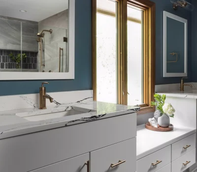 Modern bathroom features grey cabinetry, marble countertop, large mirror, brass fixtures, and a window. Decor includes a plant arrangement on a wooden tray.
