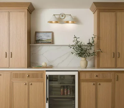 A kitchen features wooden cabinets, a marble backsplash, a mini fridge, artwork, and a vase with greenery under elegant lighting.