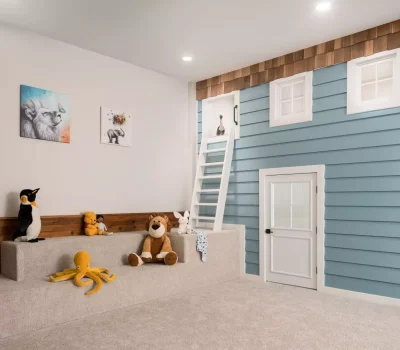 A cozy playroom with a small ladder, blue wall, stuffed animals, and animal-themed artwork on a white wall. Carpet flooring completes the space.