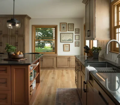 Modern kitchen with wooden cabinets, black countertops, island with plants, and wall art; large windows reveal outdoor greenery and light wood flooring.