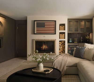 Cozy living room with American flag art, fireplace, beige sofa, and decorative shelves. Neutral tones and soft lighting create a warm atmosphere.