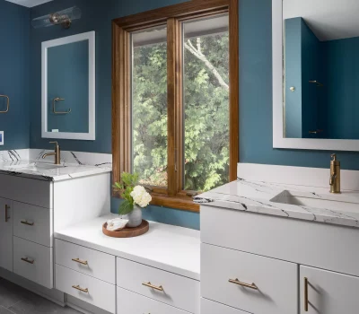Modern bathroom features marble countertops, blue walls, dual mirrors, and a central wood-trimmed window with a greenery view. Elegant gold hardware accents.