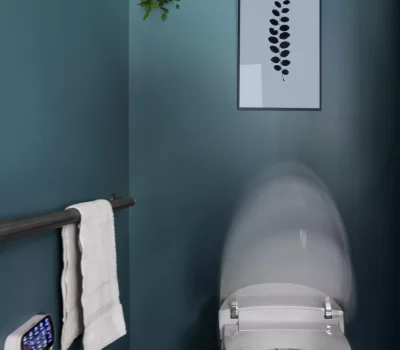 A modern automated toilet with a closing lid, teal walls, hanging plant, handrail with towel, and leaf artwork on the wall.