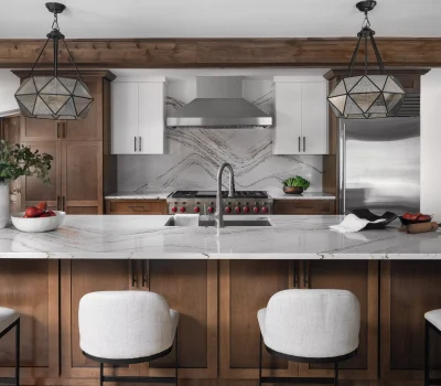 Modern kitchen with wood cabinets, marble countertops, geometric pendant lights, and stainless steel appliances. Includes a vase with greenery and red fruit.
