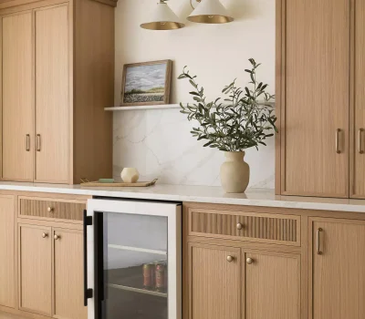 Modern kitchen scene with wooden cabinets, a small fridge, decorative plant, wall art, and dual overhead lighting, creating a cozy atmosphere.
