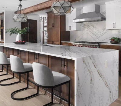 Modern kitchen with marble island, geometric pendant lights, wooden cabinetry, and upholstered chairs. Sleek stainless steel appliances and decorative green plant accents.