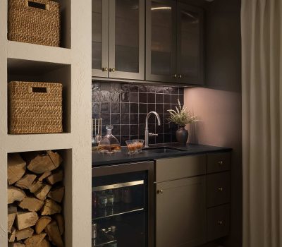 A cozy kitchen corner featuring a sink, stylish cabinetry, wood storage, glass decanter, jars, and a small refrigerator, accented by elegant lighting.