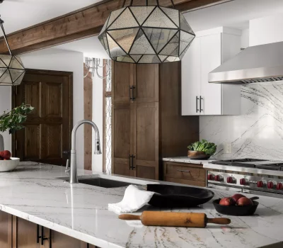 Modern kitchen features marble countertops, wooden cabinets, geometric pendant lights, and a stainless steel stove. Decor includes a bowl of fruit.
