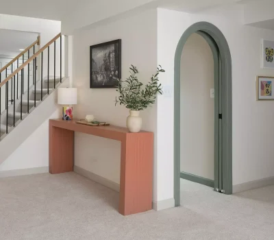 Bright interior with stairs, a wooden console, lamp, potted plant, artwork on walls, and arched doorway. Soft lighting and neutral colors.