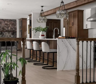Modern kitchen with marble island, wooden cabinets, geometric pendant lights, and bar stools. Plant decor and stone fireplace accentuate the cozy ambiance.