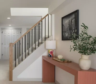 A modern interior with a staircase, red table, lamp, potted plant, and artwork depicting a historical city scene on the wall.