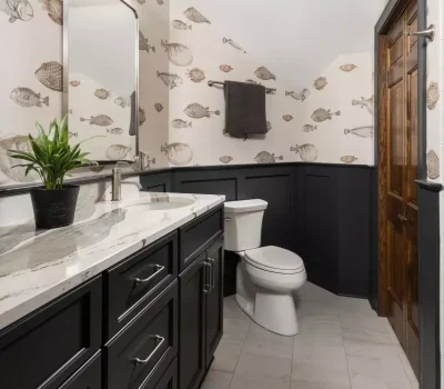 A bathroom features fish-patterned wallpaper, a marble countertop with dark cabinets, a potted plant, and a white toilet on tiled flooring.