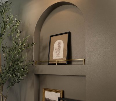 An alcove features framed artwork with a brass picture light above. Near it, a potted olive tree adds a touch of nature.