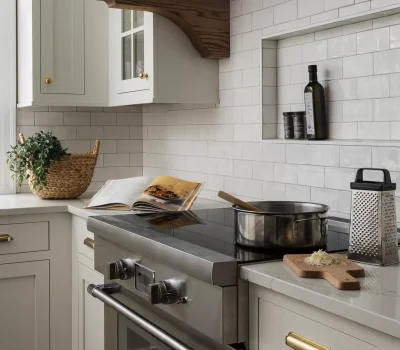 A modern kitchen features a stove, cookware, an open cookbook, olive oil, a grater, and a cutting board with cheese.