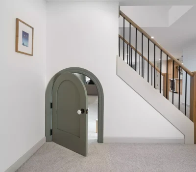A modern, carpeted hallway features a small, green arched door under a staircase, with framed artwork adorning the white wall.