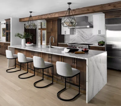 Modern kitchen with wooden cabinets, marble countertops, and stainless steel appliances. Four chairs line the island. Two geometric chandeliers hang above.