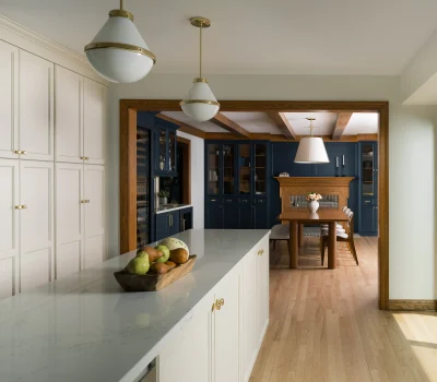 Modern kitchen with white and navy cabinets, wooden dining table, pendant lights, and hardwood flooring. Large window provides natural light.