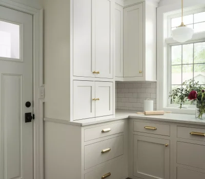 Bright kitchen corner with white cabinets, brass handles, wooden floor, and large window. A rolling pin and flowers sit on the countertop.