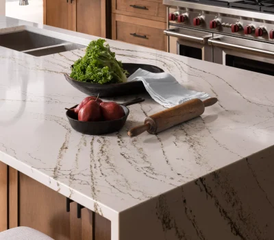 A modern kitchen features a marble countertop with a sink, rolling pin, and bowls of lettuce and pears near stainless steel appliances.