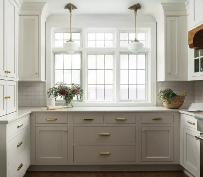 Bright kitchen with white cabinets, gold handles, and large windows. Includes flowers, a basket, and an open cookbook on the counter.