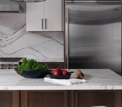 Modern kitchen with marble countertops, stainless steel fridge, geometric light fixture, and wooden cabinets. Includes chairs, greens, pears, and a rolling pin.