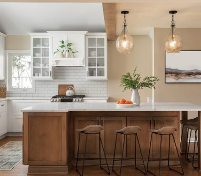Modern kitchen with white cabinets, wooden island, and bar stools. Decor includes hanging lights, potted plant, and wall art. Bright, inviting space.
