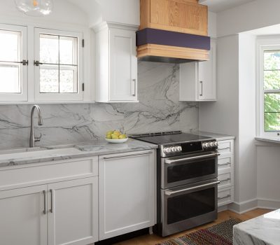 Modern kitchen with white cabinets, marble countertops, and stainless steel appliances. Bright windows illuminate the clean, sleek design, featuring a small fruit bowl.