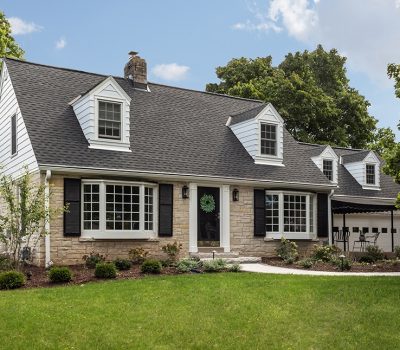 A suburban house with dormers, landscaped yard, and garage under a sunny sky, surrounded by trees and greenery.