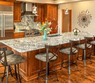 Spacious kitchen with granite island, wooden cabinetry, modern appliances, bar stools, pendant lights, and decorative wall art. No people present.